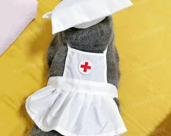 May include: A gray cat wearing a white nurse's uniform, including a cap and apron. The apron and cap feature a red cross emblem. The cat's fur is visible, and the background is a solid yellow.