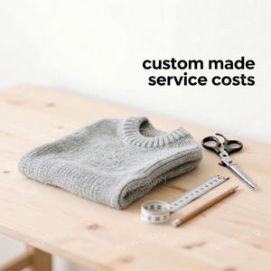May include: A folded gray knitted sweater, scissors, a measuring tape, and a pencil are arranged on a light wood surface. The text "custom made service costs" is displayed in black above the sweater.