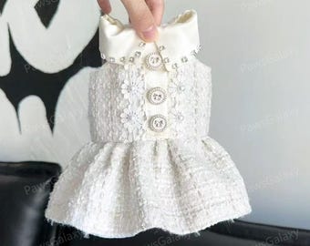 May include: A white dog dress made from textured tweed fabric. It has a satin bow collar with rhinestones and three decorative buttons. The flared skirt is complemented by floral embellishments. The dress is designed for a small dog.