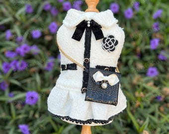 May include: White tweed dog dress with black trim and a black chain strap purse with a white bow. The dress has a black flower brooch.
