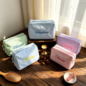 Könnte beinhalten: Fünf gestreifte Kosmetiktaschen in verschiedenen Farben: Grün, Blau, Hellblau, Rosa und Lila. Jede Tasche hat einen anderen Namen aufgestickt: Harper, Ashley, Haphine, Stacey und Stella. Die Taschen sind rechteckig mit Reißverschluss.