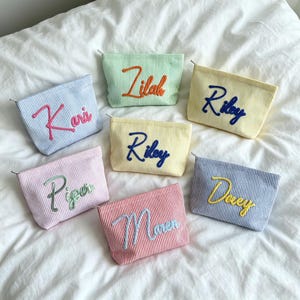 Puede incluir: Bolsas de cosméticos de pana en varios colores pastel, cada una bordada con un nombre diferente. Las bolsas presentan un patrón de rayas y un cierre de cremallera. Los nombres incluyen Kari, Lilah, Riley, Piper, Maren y Dewey.