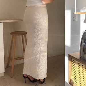 May include: A cream crochet skirt with a floral design, worn with a white vest top. The skirt is figure-hugging and ankle-length. The person is wearing black high heels.