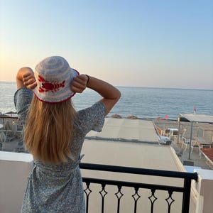 Pu&ograve; includere: Una donna indossa un cappello a secchiello a righe blu e bianche con un design di aragosta rossa. Il cappello &egrave; all'uncinetto e ha un'ampia tesa. La donna &egrave; in piedi su un balcone con vista sul mare.