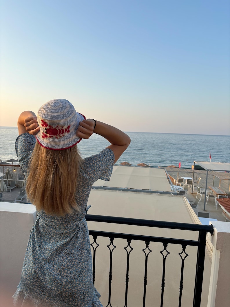 Pu&ograve; includere: Una donna indossa un cappello a secchiello a righe blu e bianche con un design di aragosta rossa. Il cappello &egrave; all'uncinetto e ha un'ampia tesa. La donna &egrave; in piedi su un balcone con vista sul mare.