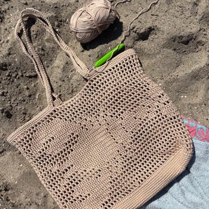 May include: A beige crocheted tote bag with long handles lies on sand, beside a ball of yarn and a green crochet hook. The open-weave design of the bag is visible, suggesting a handmade item, ideal for carrying beach essentials.
