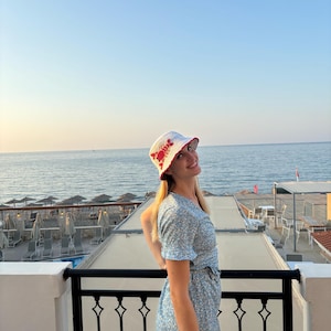 Pu&ograve; includere: Una donna indossa un cappello a secchiello bianco con un design di granchio rosso, un vestito floreale blu e sta su un balcone con vista sul mare.