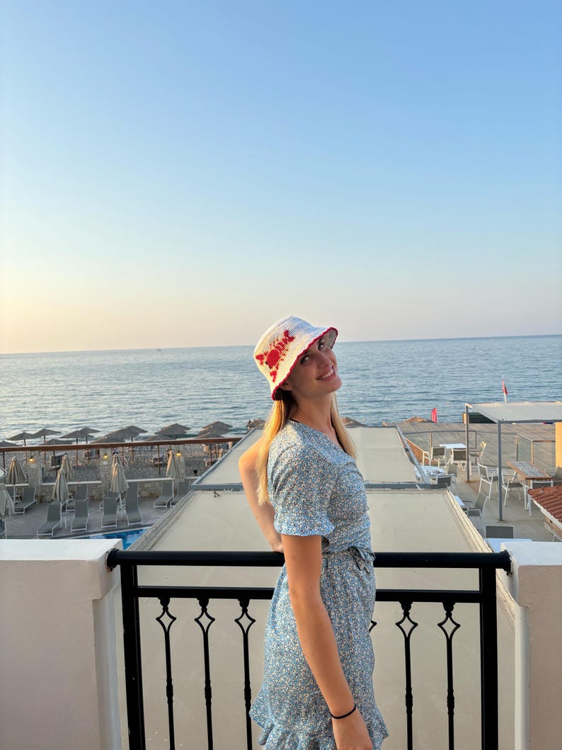 Pu&ograve; includere: Una donna indossa un cappello a secchiello bianco con un design di granchio rosso, un vestito floreale blu e sta su un balcone con vista sul mare.