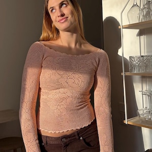 Op de afbeelding: Een perzikkleurige, gehaakte top met lange mouwen en een geschulpte halslijn en manchetten. De top heeft een gedetailleerd openwerkpatroon over de lijf en mouwen. Het model draagt een bruine broek.
