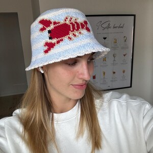 Pu&ograve; includere: Un cappello a secchiello a righe bianche e blu con un design di aragosta rossa all'uncinetto. Il cappello &egrave; indossato da una persona con i capelli lunghi e biondi.