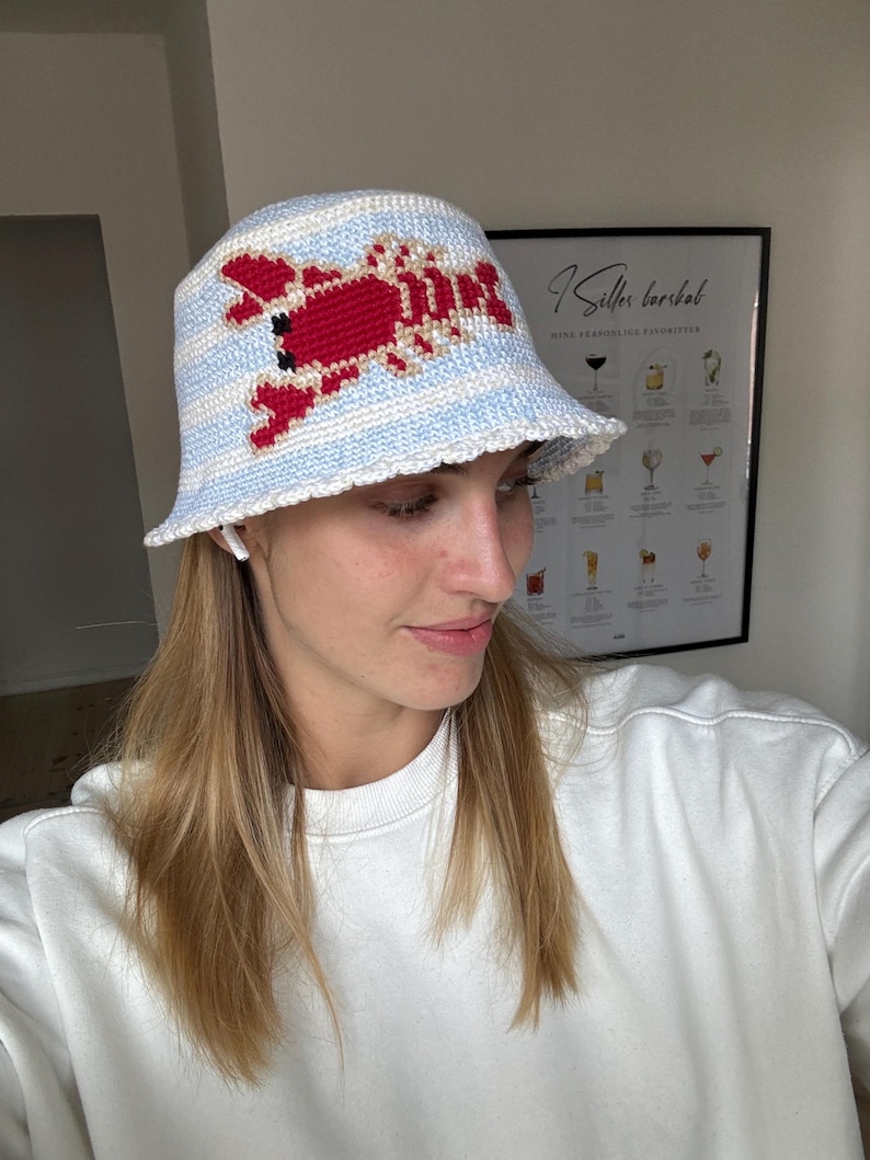 Pu&ograve; includere: Un cappello a secchiello a righe bianche e blu con un design di aragosta rossa all'uncinetto. Il cappello &egrave; indossato da una persona con i capelli lunghi e biondi.