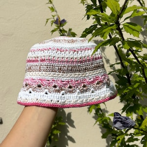 May include: A handmade crochet bucket hat in white, pink, and brown. The hat features a scalloped design and a pink brim. The hat is shown outdoors with green foliage in the background.