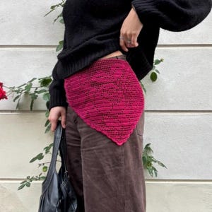 Può includere: Una gonna all'uncinetto magenta con un motivo geometrico. La gonna è di forma triangolare e indossata sopra pantaloni marroni. Un maglione nero è indossato sopra. Una borsa in pelle nera è tenuta nella mano sinistra.