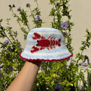 May include: A blue and white striped bucket hat with a red lobster embroidered on the front. The hat has a red trim around the brim.