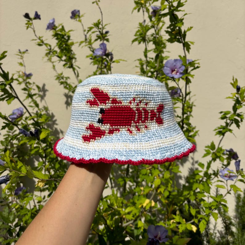 Pu&ograve; includere: Un cappello a secchiello a righe blu e bianche con un'aragosta rossa ricamata sul davanti. Il cappello ha un bordo rosso attorno al bordo.