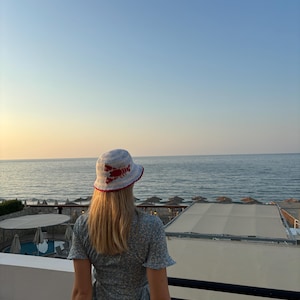 Pu&ograve; includere: Una donna con un cappello a secchiello blu e bianco con un design di aragosta rossa si trova su un balcone con vista sul mare. Indossa un vestito blu floreale.
