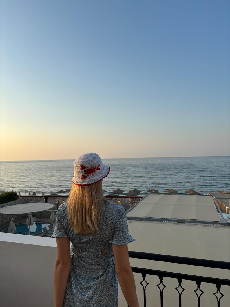 Pu&ograve; includere: Una donna con un cappello a secchiello blu e bianco con un design di aragosta rossa si trova su un balcone con vista sul mare. Indossa un vestito blu floreale.