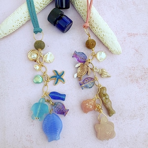 May include: Two handmade sea-themed car charms. One charm has a teal suede cord, the other a pink cord. Each charm features gold-toned chains and a variety of ocean-themed beads, including starfish, shells, and fish. The charms are on a pink background.