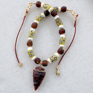 Peut inclure: Un collier avec un pendentif en forme de pointe de flèche en pierre brun foncé suspendu à un cordon. Le collier alterne des perles rondes brun foncé, des entretoises cylindriques blanches et des perles cylindriques jaunes à motifs d'yeux.