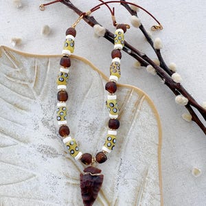 May include: A necklace featuring a dark brown arrowhead pendant wrapped in gold-toned wire. The strand consists of alternating round dark brown beads, cylindrical white beads, and yellow cylindrical beads with black and white circular patterns.