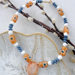 May include: A necklace featuring cylindrical orange beads with white and teal floral patterns, interspersed with round white beads and faceted slate blue beads. A large, irregular, translucent orange stone pendant hangs from the centre.