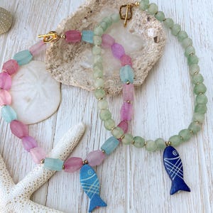 May include: Two beaded necklaces featuring frosted glass beads in shades of pink, light blue, and pale green. Each necklace includes a gold-toned toggle clasp and a flat, carved fish-shaped pendant in either bright blue or dark navy blue.