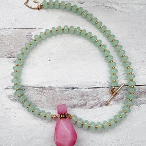 May include: A delicate necklace featuring light green beads with gold accents. A pink, faceted pendant hangs from the center. The necklace has a gold heart-shaped clasp and a gold bar clasp.