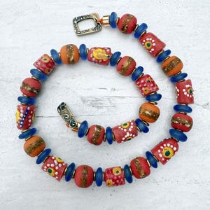Peut inclure: Un collier composé d'une série de perles cylindriques et sphériques dans des tons de rouge, orange et bleu. Les perles rouges sont ornées de motifs peints en blanc et jaune. De petites perles disques bleues sont intercalées entre elles.