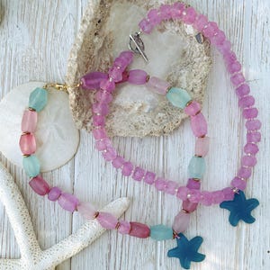 Op de afbeelding: Twee kettingen met gematteerde kralen in roze, paarse en lichtblauwe tinten, geaccentueerd met kleine metalen tussenstukjes. Elke ketting heeft een donkerblauwe hanger in de vorm van een zeester en een zilverkleurige knevelsluiting.
