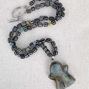 May include: A necklace featuring a ghost-shaped pendant carved from iridescent labradorite stone. The strand consists of dark brown barrel-shaped beads with white vertical stripes, accented by small blue and yellow glass beads and a silver toggle clasp.