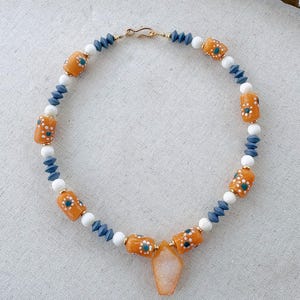 Könnte beinhalten: Eine Perlenkette mit zylindrischen orangefarbenen Perlen mit weißen und türkisfarbenen Blumenmustern, durchsetzt mit kleinen weißen runden Perlen und facettierten schieferblauen Perlen. Ein durchsichtiger, hellorangefarbener, sargförmiger Anhänger hängt in der Mitte.