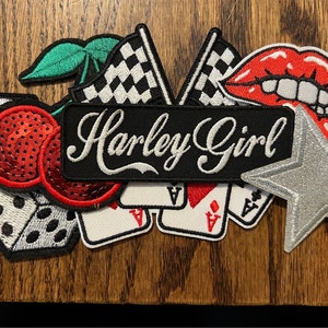 Könnte beinhalten: Eine Sammlung gestickter Aufnäher mit verschiedenen Designs. Enthält einen schwarzen Aufnäher mit "Harley Girl" in weißer Schrift, einen silbernen Stern, rote Lippen, Würfel, Kirschen, Spielkarten und Schachflaggen.
