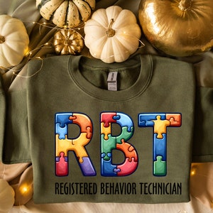 May include: An olive green sweatshirt with the letters "RBT" in a colorful puzzle design. Below the letters, the text reads "REGISTERED BEHAVIOR TECHNICIAN". The sweatshirt is surrounded by decorative pumpkins.