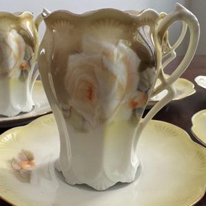 May include: A set of porcelain teacups and saucers featuring a cream and pale yellow color palette with hand-painted peach-colored rose motifs. Each cup has a scalloped rim, a curved handle, and a flared base, while the matching saucers have a scalloped edge.