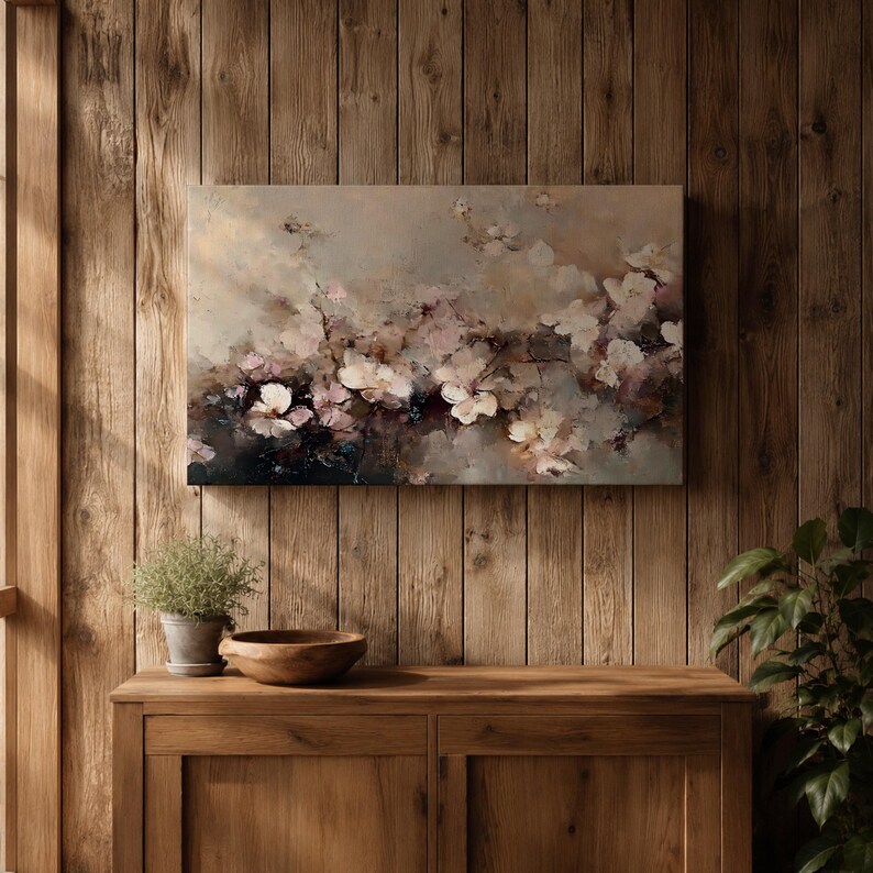 Puede incluir: Una pintura floral abstracta en tonos apagados de beige, rosa y blanco cuelga de una pared de madera. La obra de arte representa delicadas flores y ramas, creando una est&eacute;tica suave y on&iacute;rica. Debajo, un mueble de madera con una planta y un cuenco.