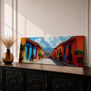 Panoramic cityscape painting showing a vibrant Mexican street lined with colorful colonial houses in bright shades of blue, red, orange, and yellow, with potted plants and balconies along a cobblestone road leading toward distant hills beneath a partly cloudy sky.