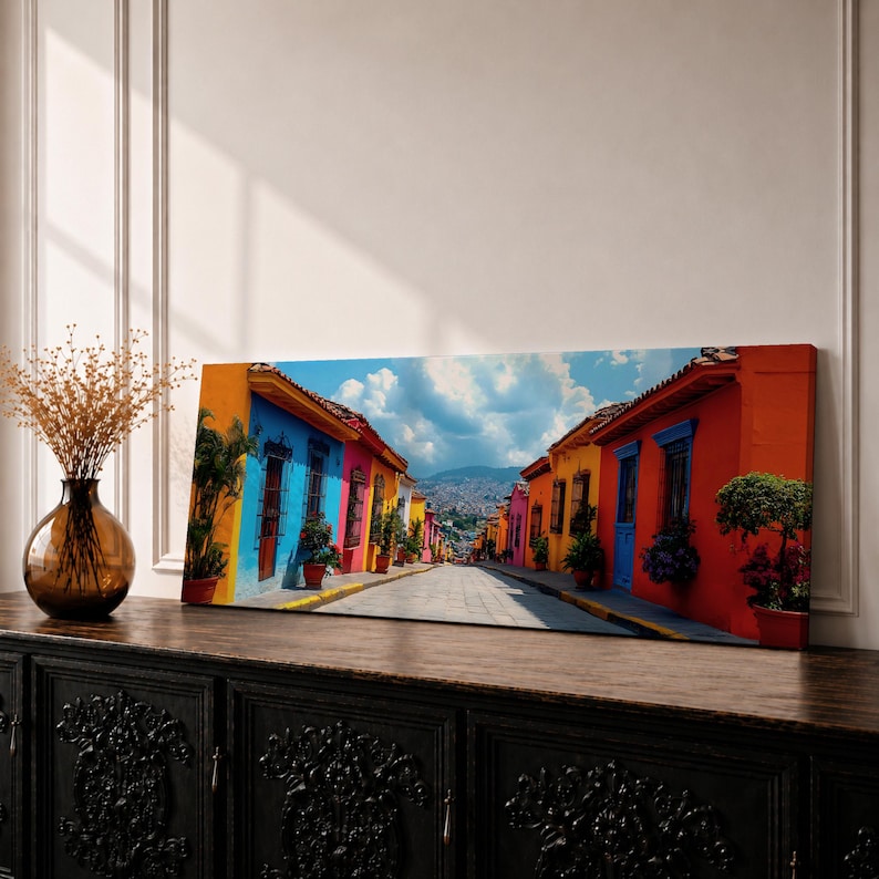 Panoramic cityscape painting showing a vibrant Mexican street lined with colorful colonial houses in bright shades of blue, red, orange, and yellow, with potted plants and balconies along a cobblestone road leading toward distant hills beneath a partly cloudy sky.