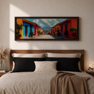 Puede incluir: Un cuadro panor&aacute;mico enmarcado que muestra una calle estrecha flanqueada por edificios de colores vibrantes en tonos naranja, azul, amarillo y rosa. La escena incluye plantas en macetas, puertas de madera oscura y una monta&ntilde;a lejana.