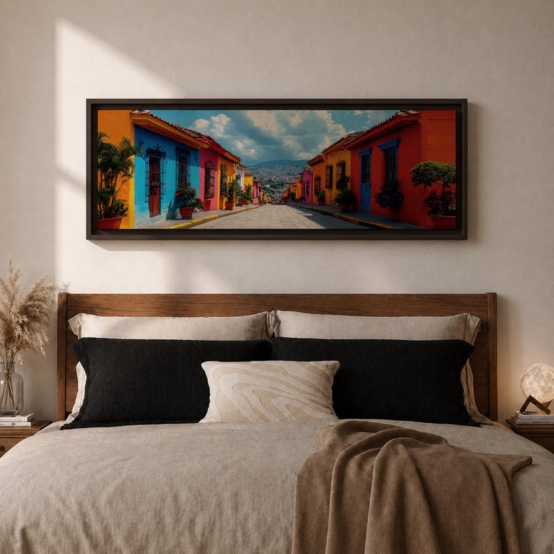Puede incluir: Un cuadro panor&aacute;mico enmarcado que muestra una calle estrecha flanqueada por edificios de colores vibrantes en tonos naranja, azul, amarillo y rosa. La escena incluye plantas en macetas, puertas de madera oscura y una monta&ntilde;a lejana.