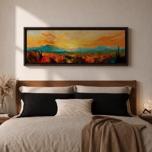 Panoramic desert sunset painting showing distant layered mountains beneath a glowing sun with sweeping orange and gold skies. Thick textured brushstrokes form desert plants and rocky terrain in the foreground while teal and blue ridges fade into the horizon, creating a wide southwestern landscape composition.