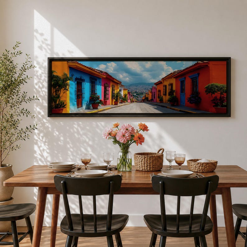 Puede incluir: Un cuadro panor&aacute;mico que muestra una escena callejera con edificios de colores azul, amarillo y naranja bajo un cielo nublado, colgado sobre una mesa de comedor de madera con sillas negras, un jarr&oacute;n de cristal con flores y cestas de mimbre.