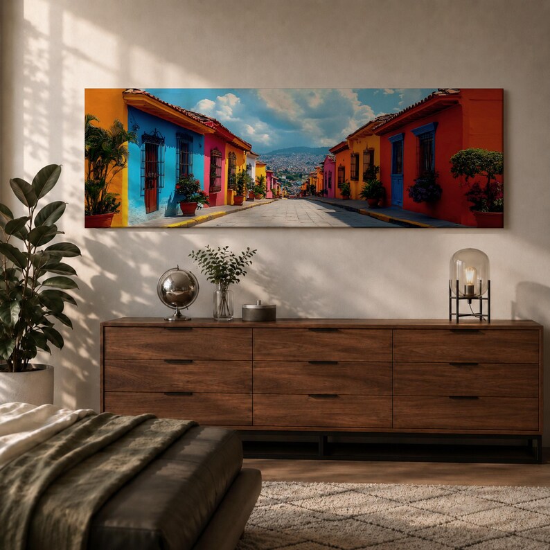 Puede incluir: Una impresi&oacute;n de arte mural panor&aacute;mico que representa una escena callejera vibrante con edificios coloridos en tonos azules, amarillos y rojos, que conducen hacia una monta&ntilde;a lejana bajo un cielo brillante con nubes, en formato rectangular.