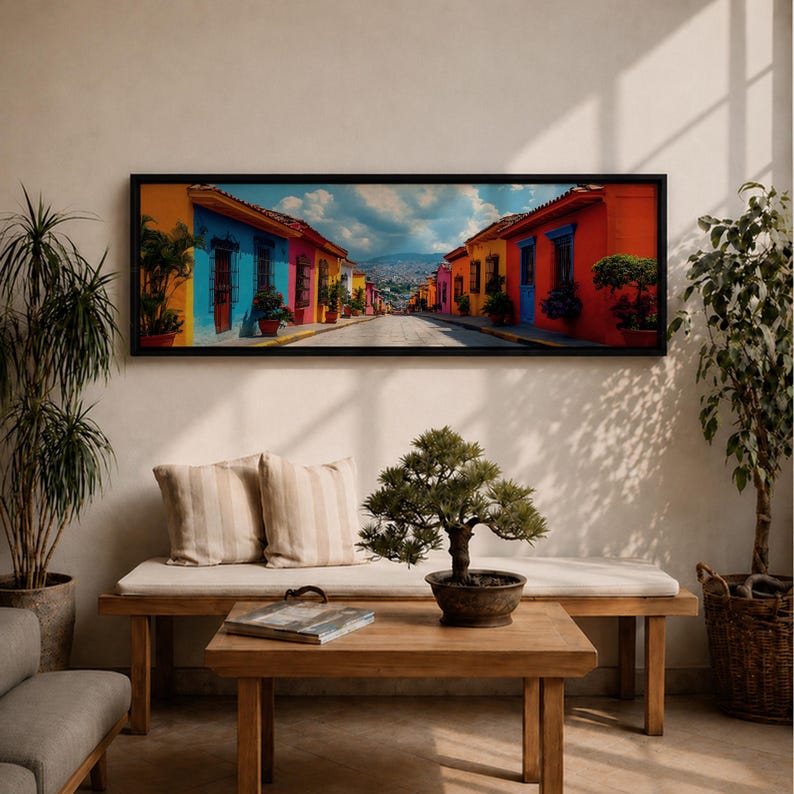 Puede incluir: Una impresi&oacute;n de arte mural panor&aacute;mico en un marco negro que representa una escena callejera con edificios de colores vibrantes azul, amarillo, naranja y rojo bajo un cielo nublado. La obra se exhibe sobre un banco de madera con dos cojines beige.