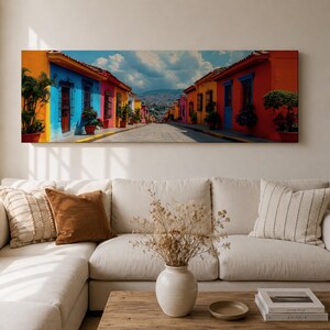 Puede incluir: Un cuadro panor&aacute;mico que representa una calle estrecha bordeada de edificios de colores vibrantes en tonos naranja, azul, amarillo y rojo, que conduce hacia monta&ntilde;as lejanas bajo un cielo azul nublado, expuesto sobre un sof&aacute; beige.