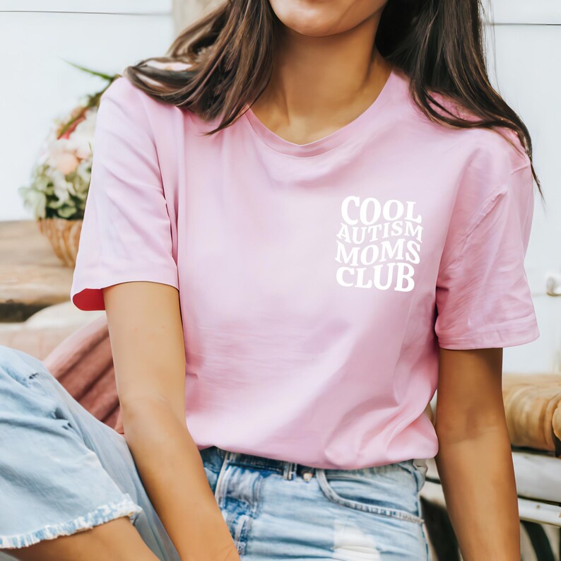 Może przedstawiać: R&oacute;żowa koszulka z napisem "COOL AUTISM MOMS CLUB" w kolorze białym.