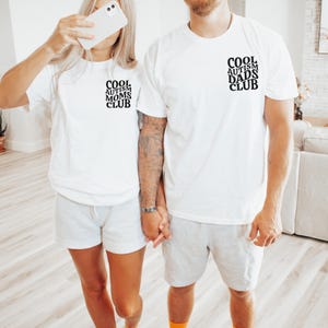 Może przedstawiać: Para w białych koszulkach z nadrukiem "Cool Autism Moms Club" i "Cool Autism Dads Club". Trzymają się za ręce i uśmiechają.