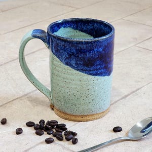 May include: A handmade ceramic mug with a blue and green glaze. The mug has a large handle and a speckled finish. Coffee beans and a spoon are next to the mug. The mug is approximately 5 inches tall.