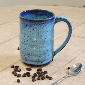 May include: A tall, handmade ceramic mug with a blue and turquoise glaze. The mug has a large handle and a textured surface. Coffee beans and a spoon are next to the mug.