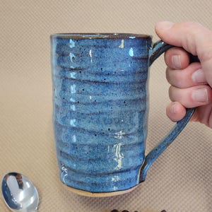 May include: A tall, cylindrical ceramic mug with a textured, rippled surface and a speckled blue glaze. The mug features a sturdy handle on the right side and a smooth, light-colored base, showcasing a handcrafted appearance.