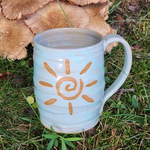 May include: A light blue ceramic mug with a ribbed, cylindrical body and a single handle. The exterior features a centered, hand-painted golden-brown sun design with a spiral center and radiating rays. The glaze has a smooth, glossy finish.